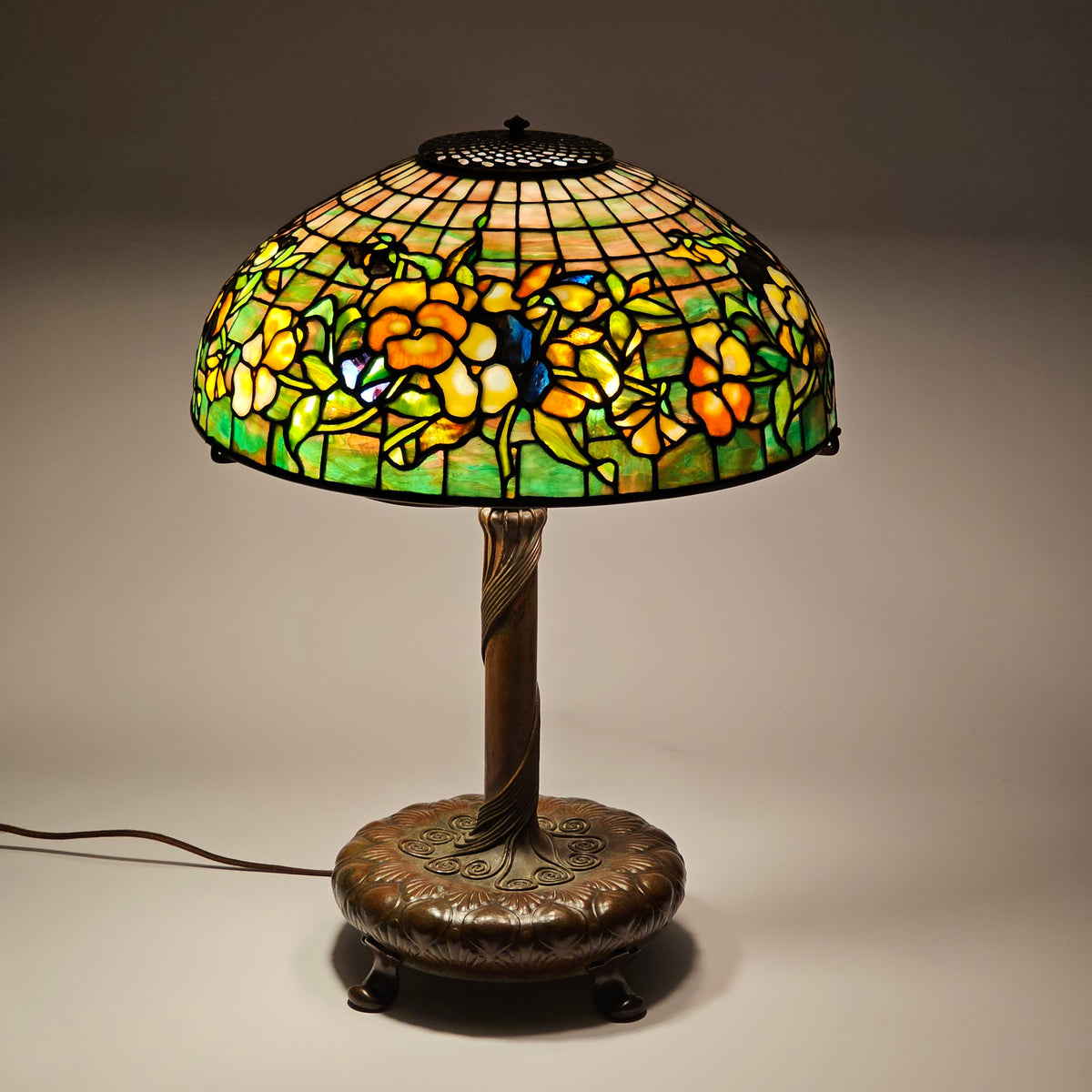 Tiffany glass table lamp with a floral design in yellow, orange, and green, mounted on a round bronze wall plate with a visible power cord against a neutral wall.